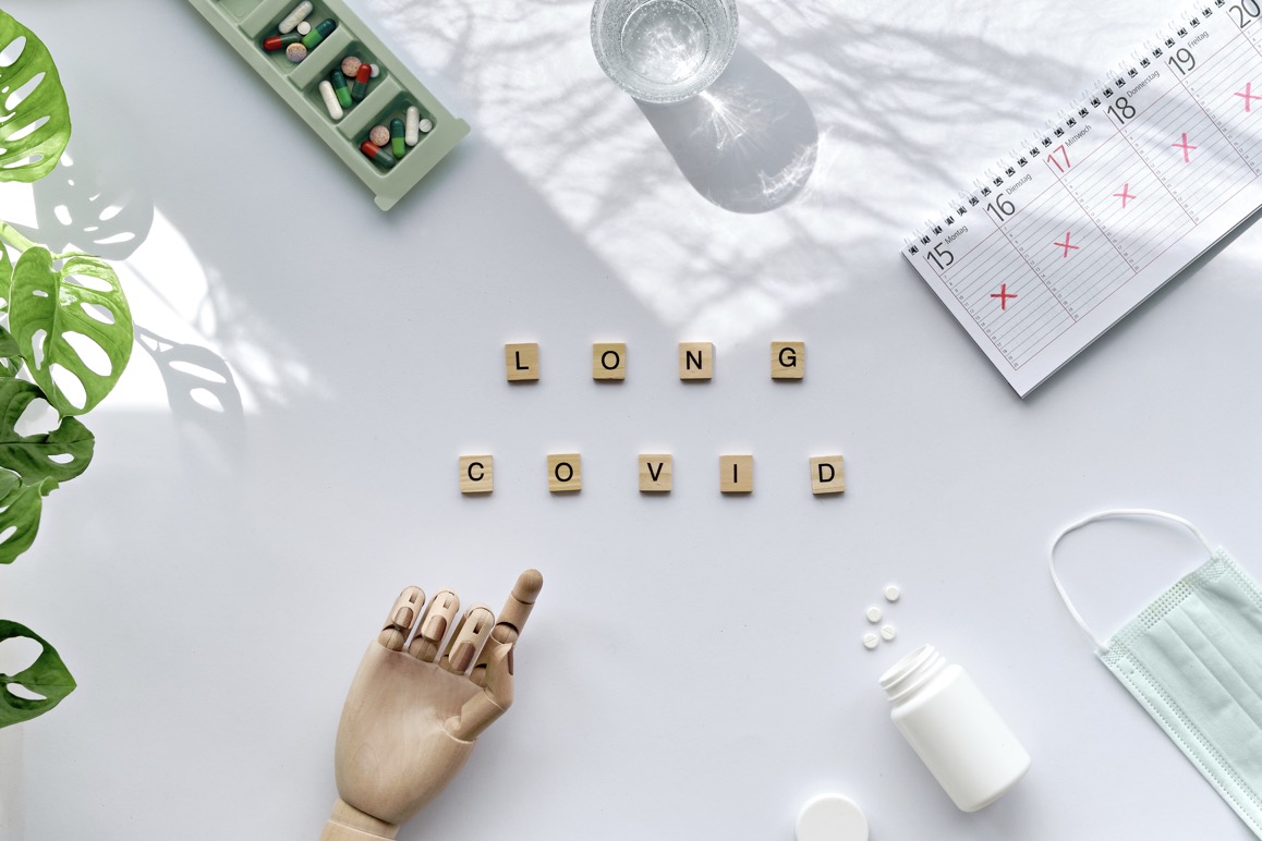 Text Long Covid, wooden letters. Wooden model hand points at weekly planner, German text means days of the week. Cancellation, change of plans, chronic covid illness. Off white table, monstera leaves.